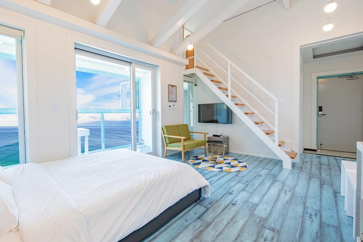 A bedroom with a large bed, a TV, a chair, and a staircase leading to an upper deck with a view of the ocean.