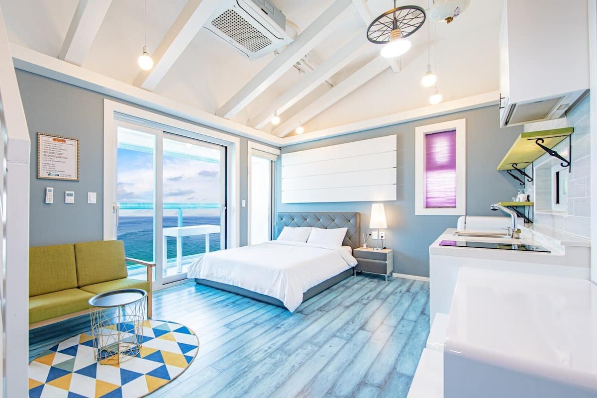A modern hotel room with a large bed, a green sofa, a round coffee table, and a view of the ocean through the balcony doors.