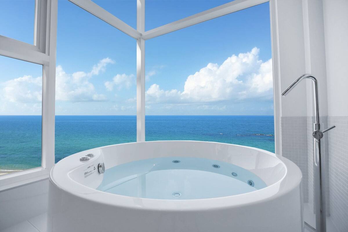 A modern bathroom with a large bathtub, a shower, and a view of the ocean through large windows.