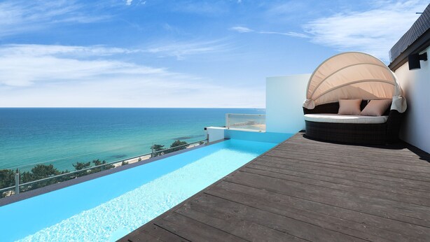 Una terraza de madera con piscina y vistas al mar, un área de estar cubierta y cielo despejado.