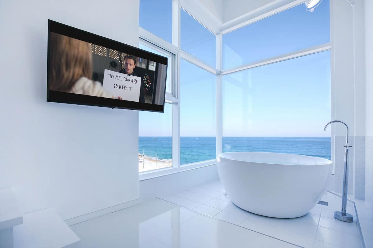 A modern bathroom with a large bathtub, a TV mounted on the wall, and a view of the ocean through large windows.