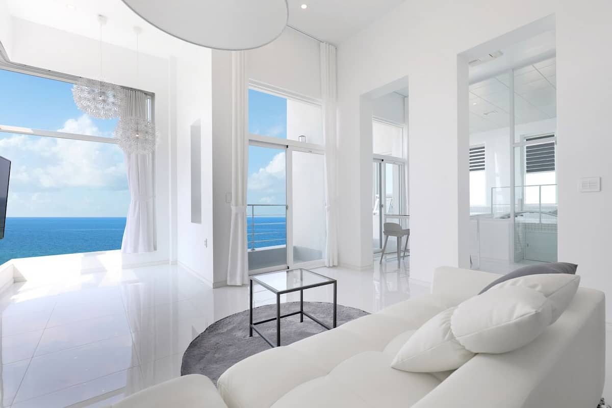 A modern living room with a white sofa, a glass coffee table, and a view of the ocean.