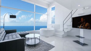 Un moderno salón con un ventanal grande que da al océano, un sofá, una mesa de centro redonda, una bañera blanca y una escalera.