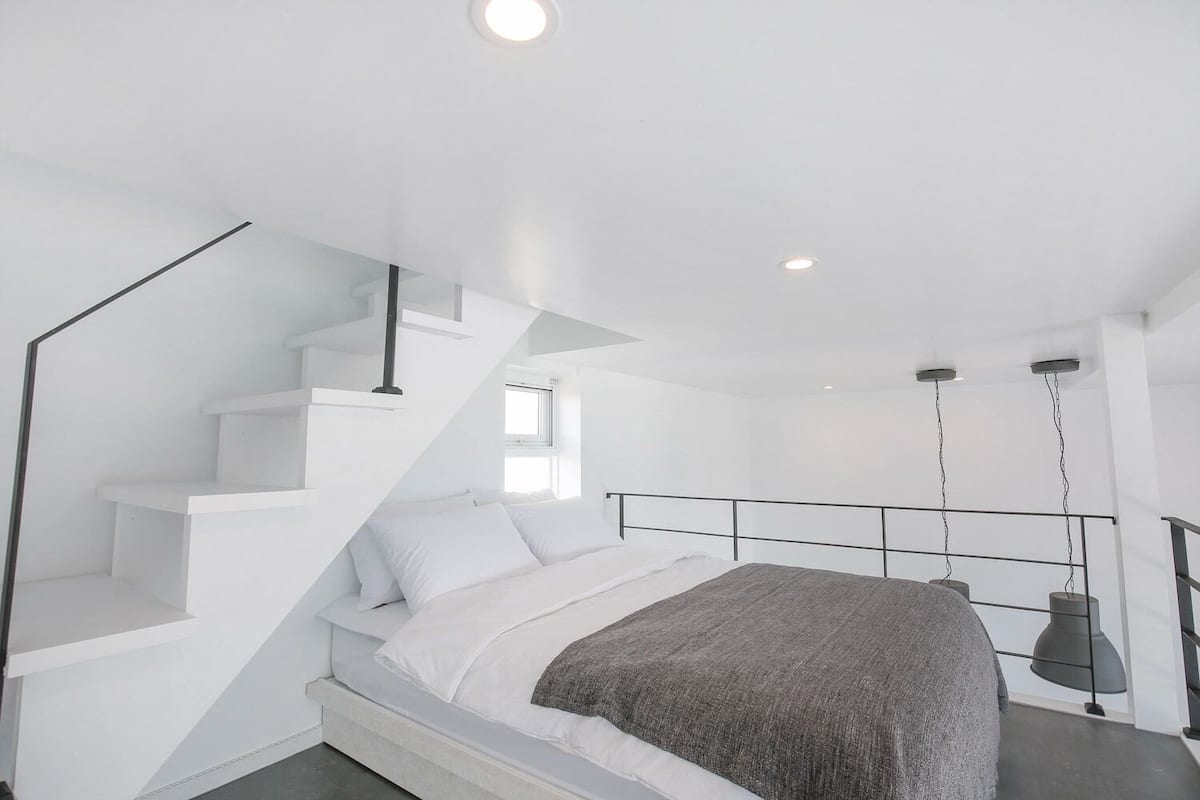 A modern bedroom with a white staircase, a bed with white and gray bedding, and a hanging light fixture.