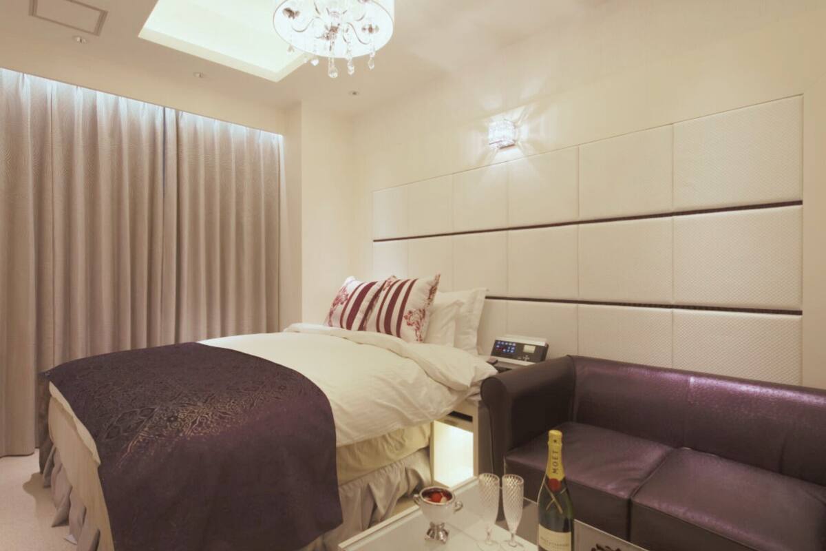 A hotel room with a bed, a purple sofa, a minibar, and a large window with curtains.