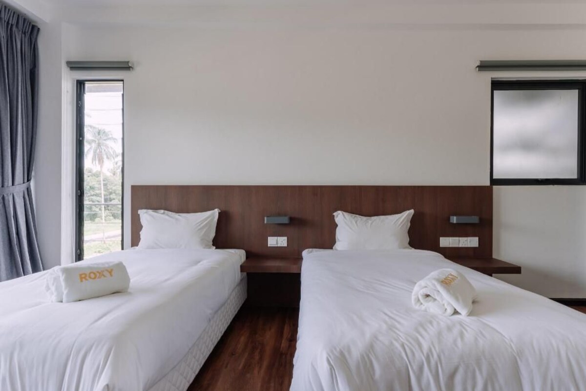 A hotel room with two beds, a wooden headboard, bedside tables, and a large window with curtains.