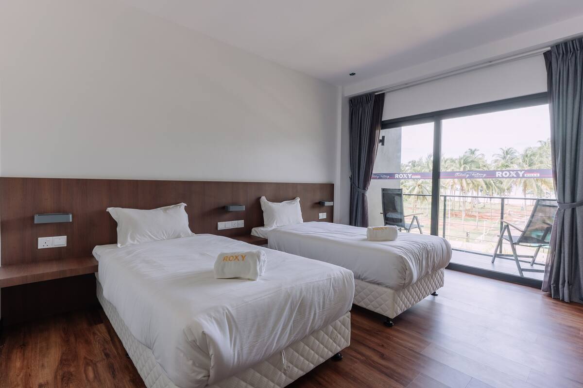 A hotel room with two beds, a wooden headboard, a large window with a view, and a balcony with chairs.