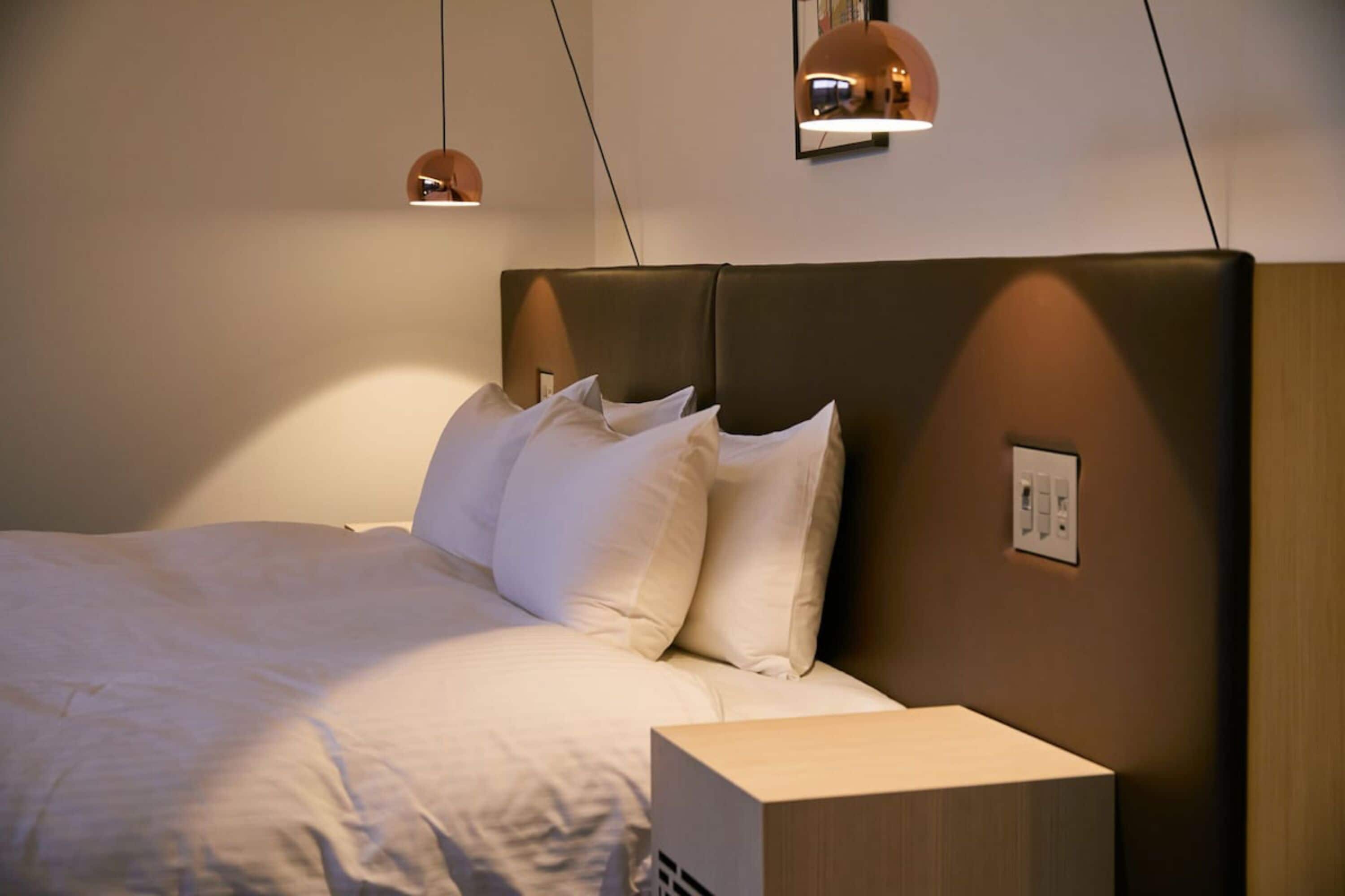 A neatly made bed with white linens and a wooden headboard. There are two pendant lights above the bed, and a small bedside table with a lamp.
