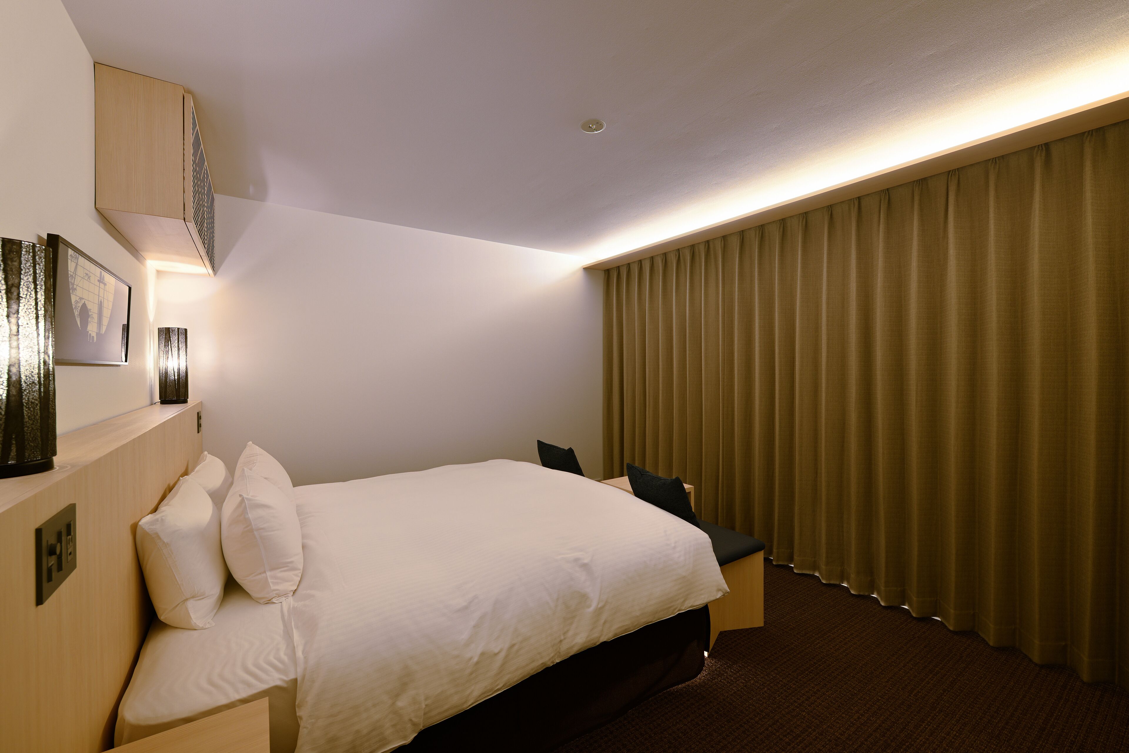 A hotel room with a large bed, a bedside table, a wall-mounted lamp, and a window with curtains.