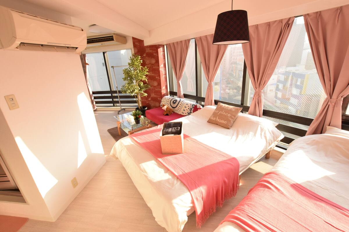 Quarto de hotel moderno com uma cama grande, uma mesinha, um vaso de planta e vista da cidade por grandes janelas.