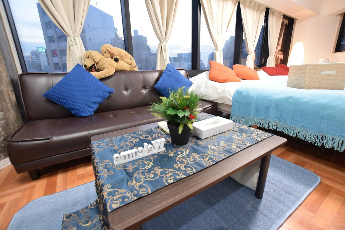 Quarto de hotel moderno com sofá de couro, cama com edredom azul e vista para a paisagem urbana através de grandes janelas.
