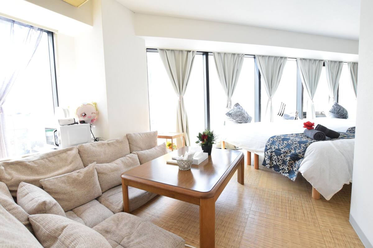 Quarto de hotel moderno com sofá, mesa de madeira, cama com colcha estampada e janelas grandes com cortinas.