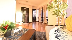 A spacious living room with a wooden floor, a large window with curtains, a potted plant, and a sofa with a patterned throw. - Balcone Uchikyuhoji (Osaka)