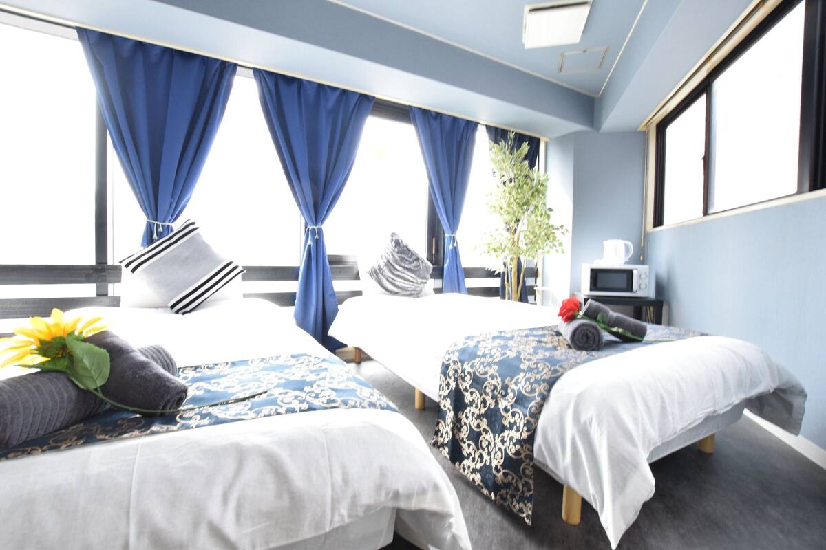 Quarto de hotel com duas camas, uma mesinha com chaleira e janela com cortinas.