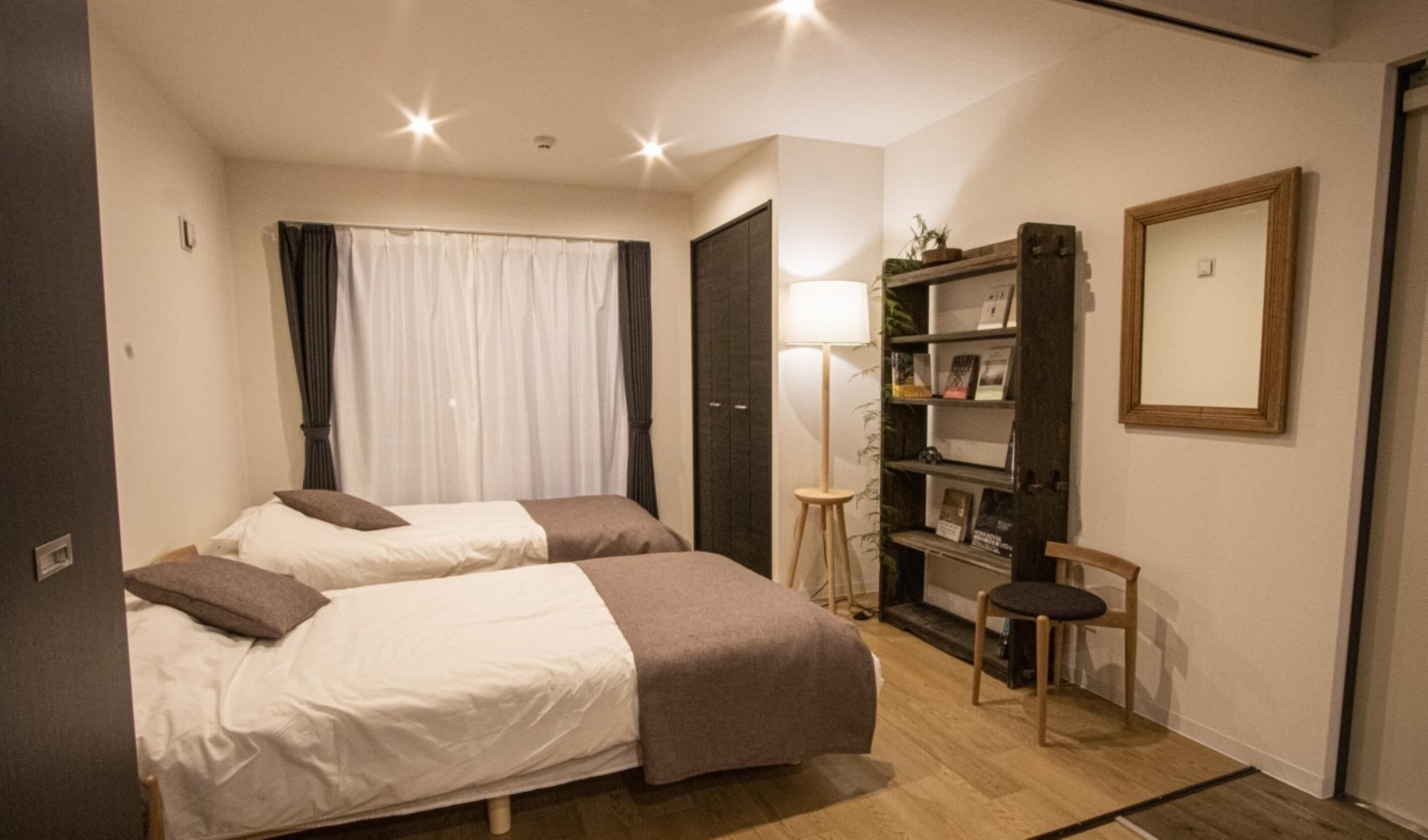 A bedroom with two beds, a bedside table, a lamp, a bookshelf, and a mirror.