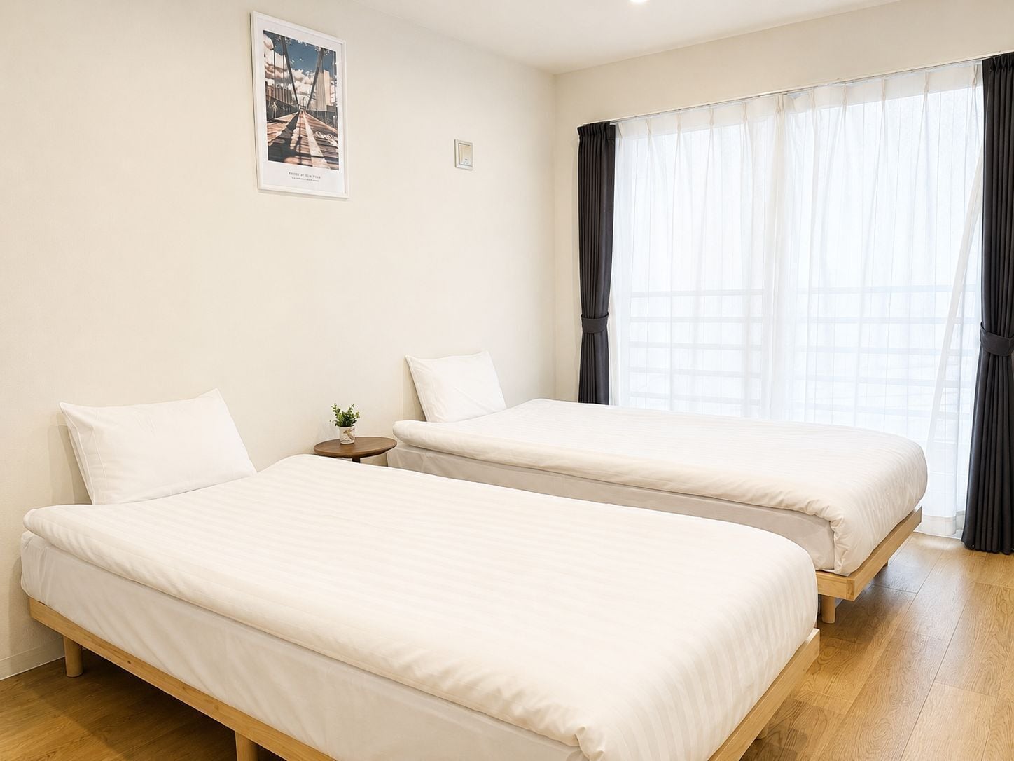 A hotel room with two single beds, a small bedside table with a plant, a framed picture on the wall, and a large window with curtains.