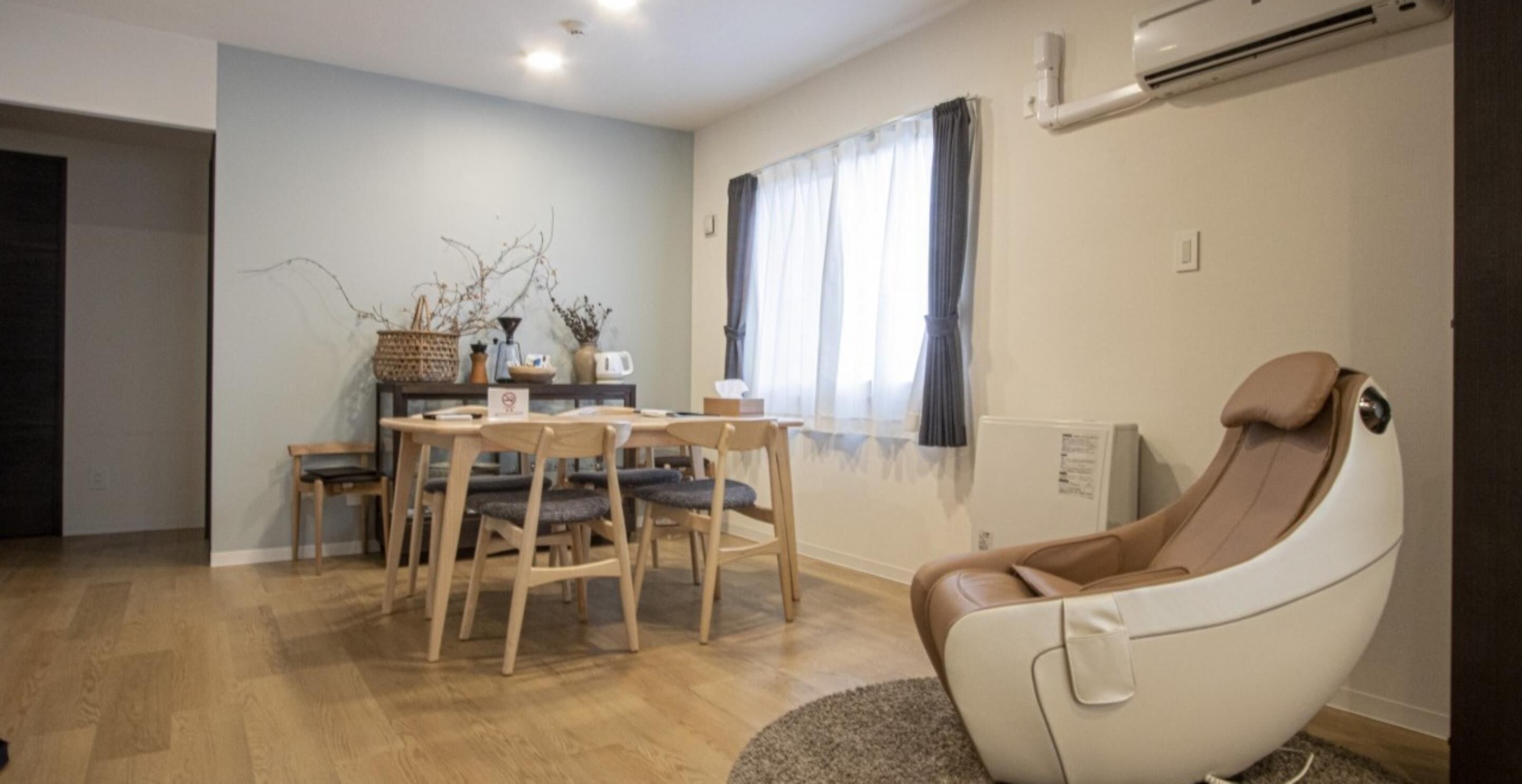 A modern living room with a dining area, a massage chair, and a window with curtains.