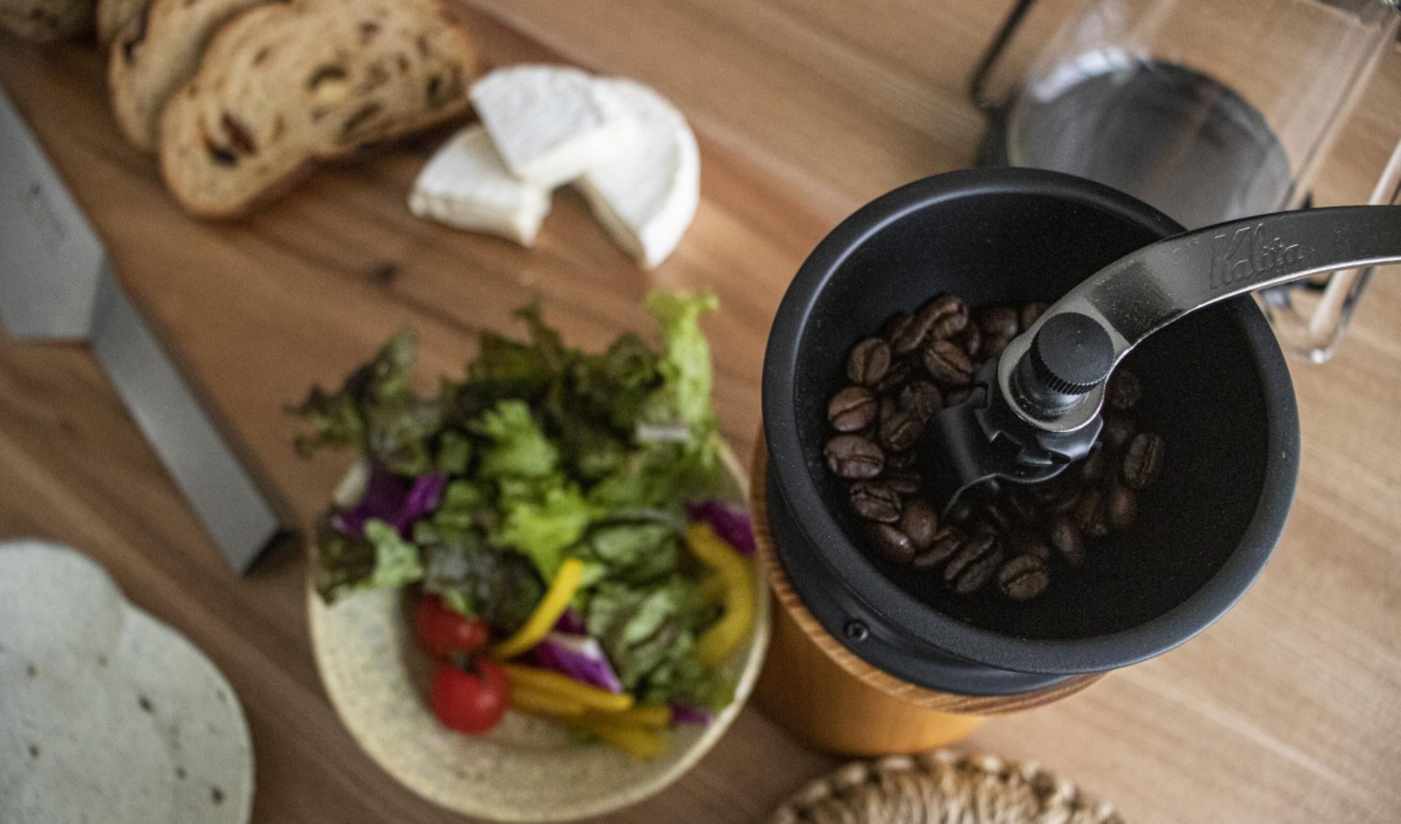 A coffee grinder with coffee beans, a plate of salad, and a loaf of bread on a wooden table.