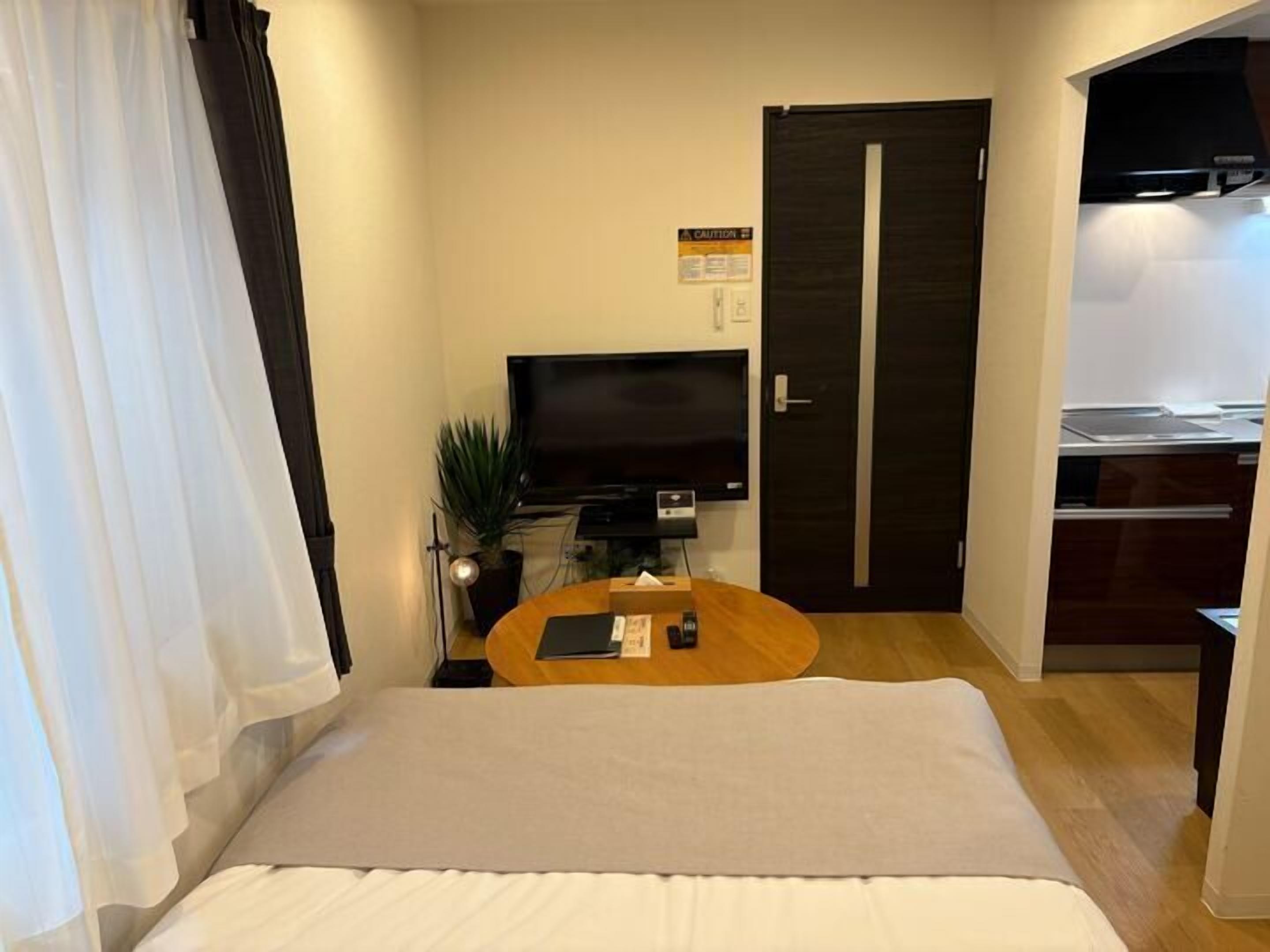 A hotel room with a bed, a television, a small table with a remote control, a potted plant, and a door leading to a kitchen area.