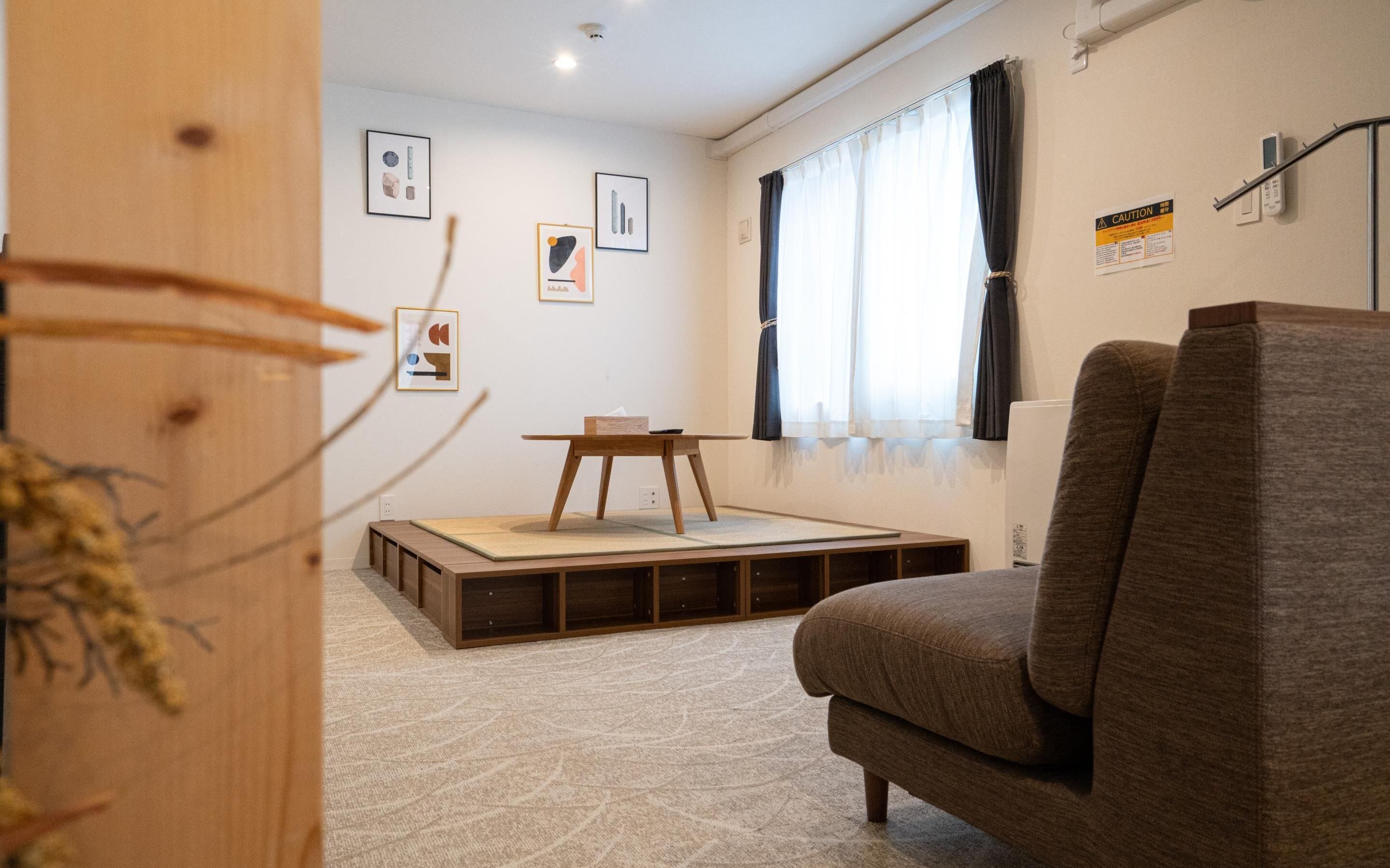 Un salon moderne avec un canapé, une petite table et un tatami. Des tableaux encadrés ornent le mur, et un panneau d’avertissement y est également affiché.