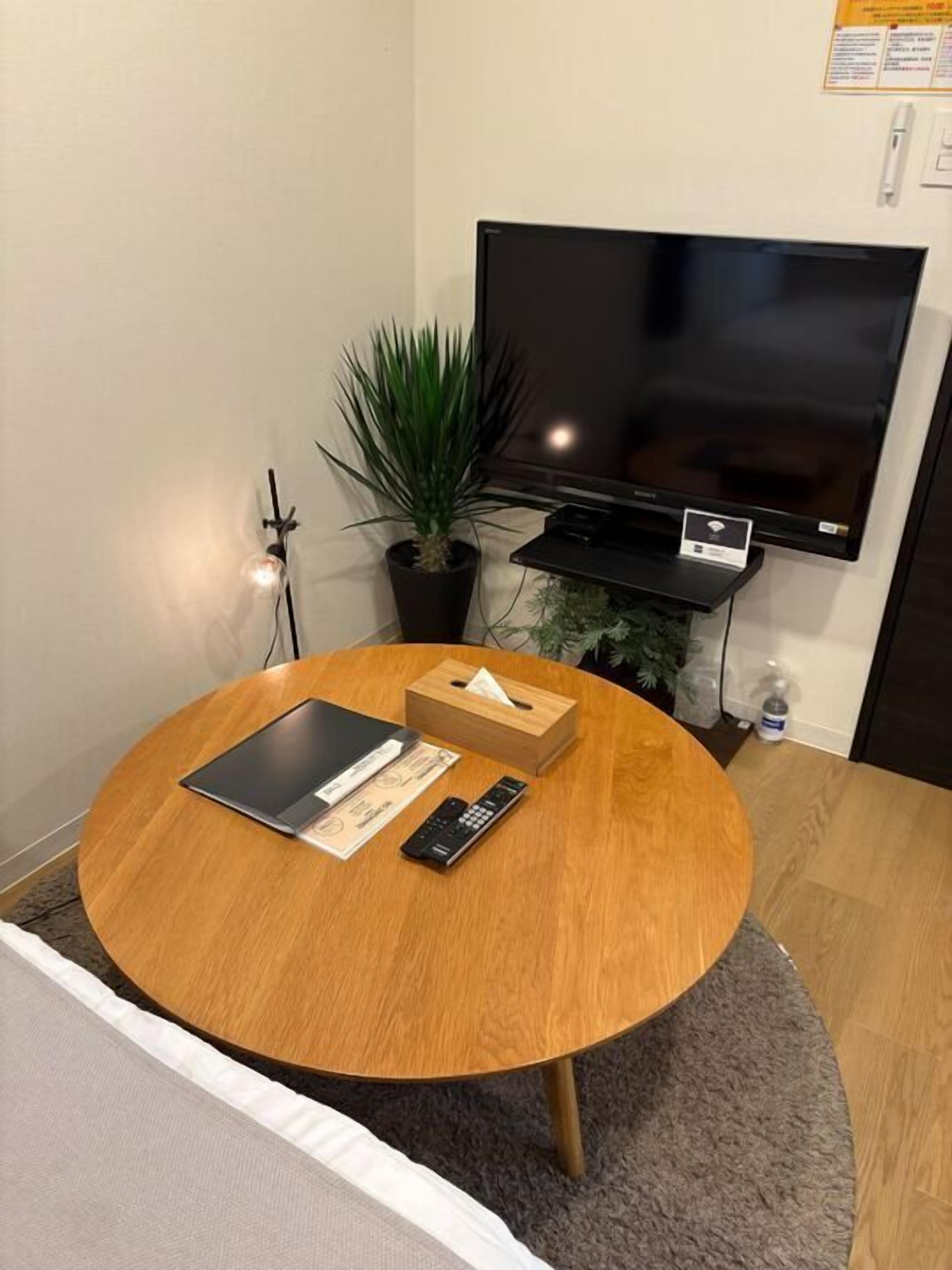 A round wooden table with a remote control, a tissue box, and a tablet. A flat-screen TV is mounted on the wall above the table.