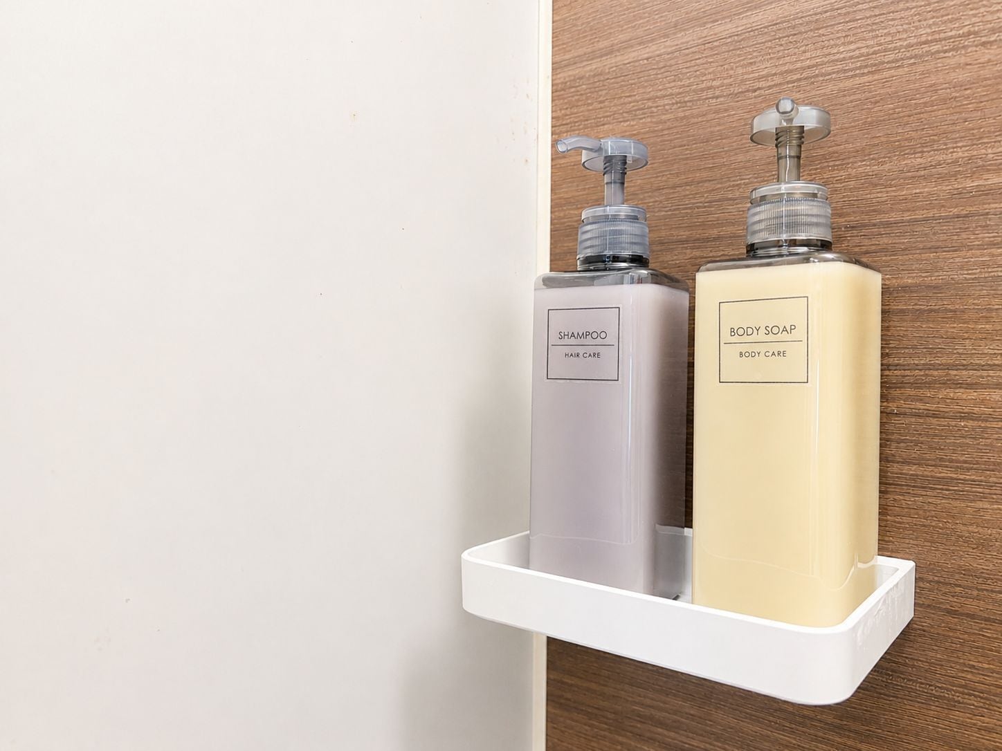 Deux bouteilles de shampoing et de savon pour le corps sont posées sur une étagère blanche, devant un mur en bois.