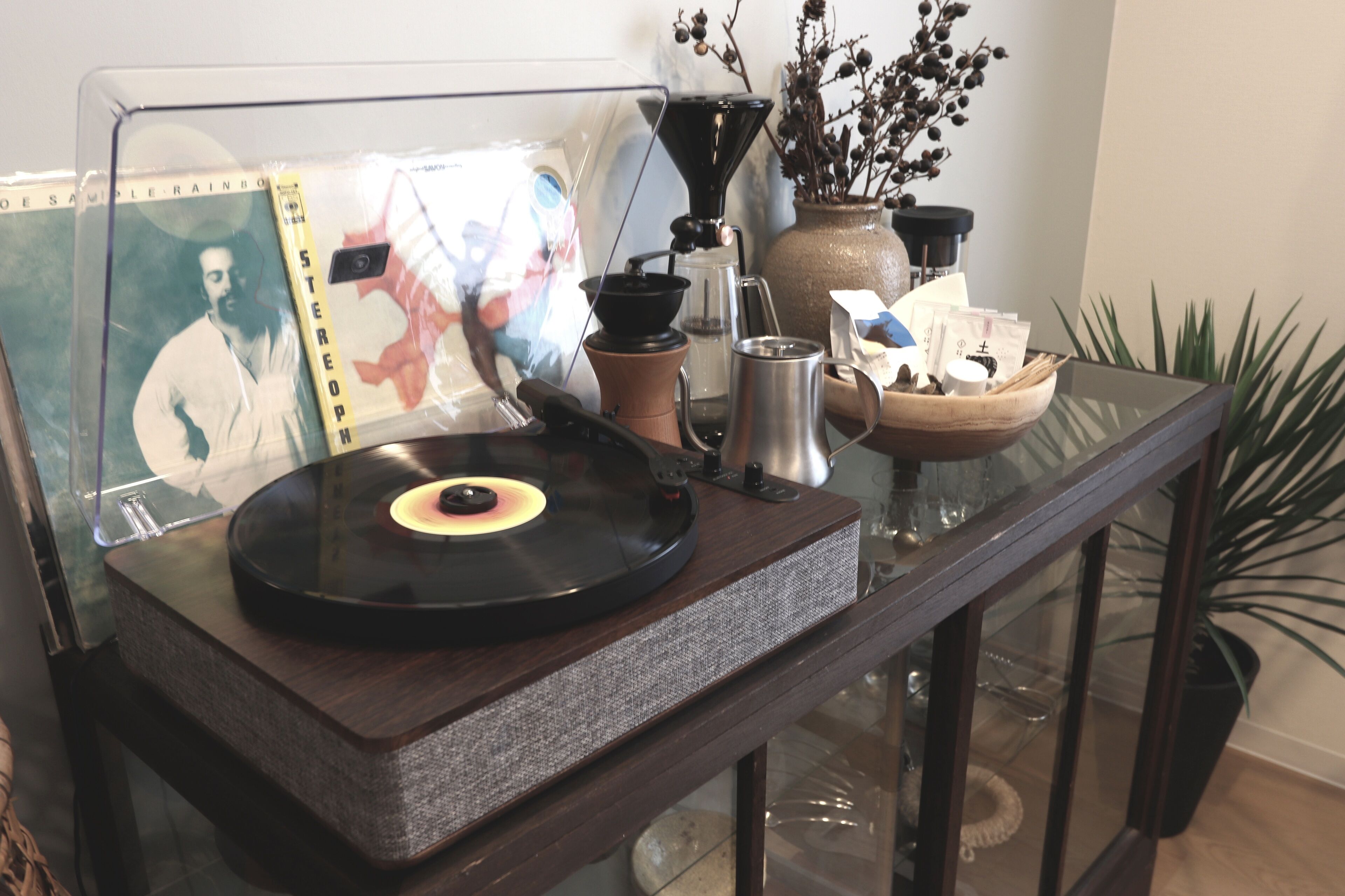 A vinyl record player on a glass table with a coffee maker, a vase with dried branches, and a framed picture.