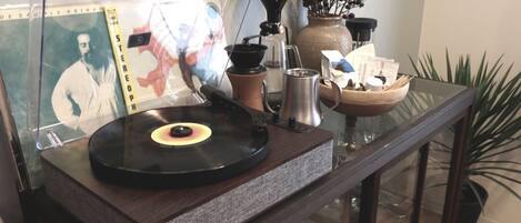 A vinyl record player on a glass table with a coffee maker, a vase with dried branches, and a framed picture.