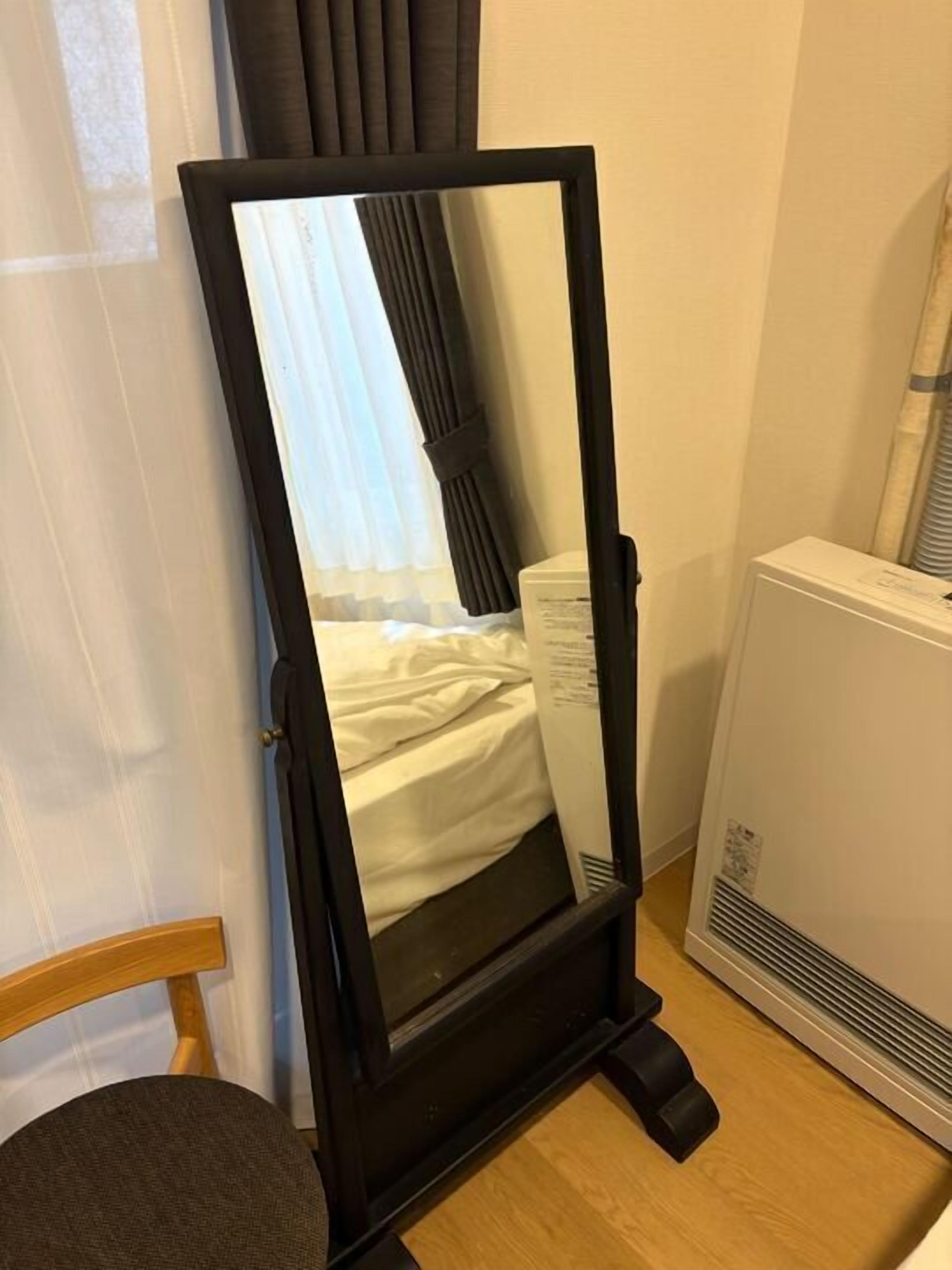 Una habitación con cama, un espejo de pie, una silla y un calefactor.