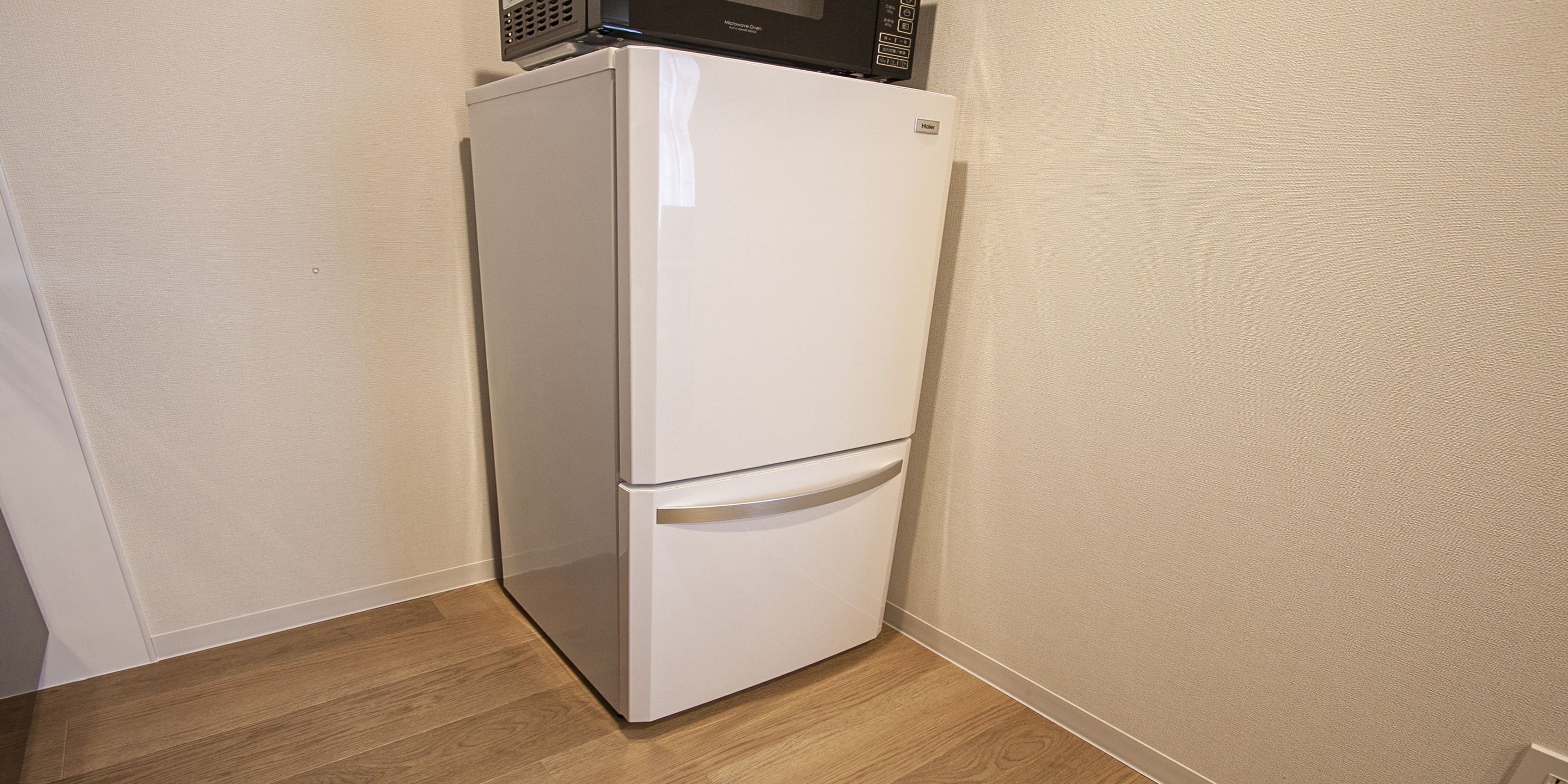 A white refrigerator with a microwave on top, located in a corner of a room with wooden flooring and beige walls.