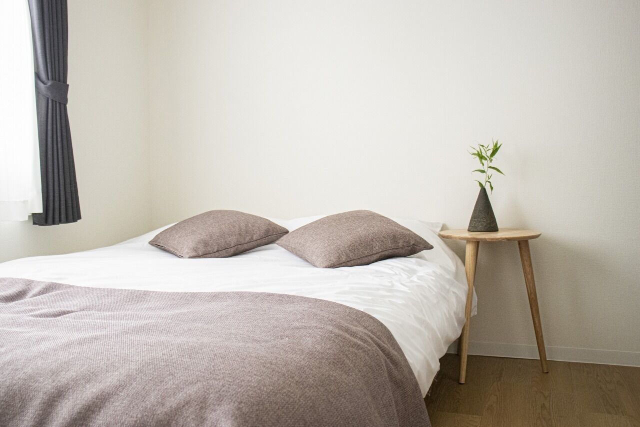 A neatly made bed with a grey blanket and two pillows, a small wooden side table with a plant, and a window with grey curtains.