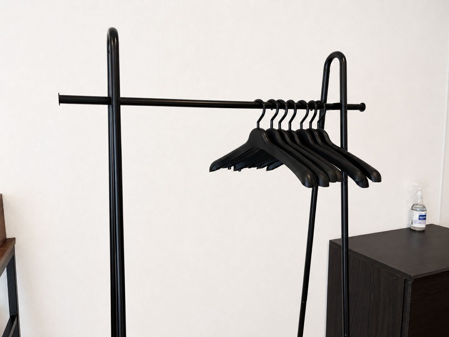 A black metal rack with multiple hangers against a white wall, a small wooden table, and a bottle of hand sanitizer on a dark wooden surface.