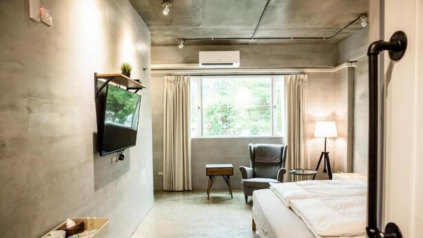 A hotel room with a bed, a TV mounted on the wall, a small table, a chair, a lamp, and a window with curtains. - Genuinn (Taitung)