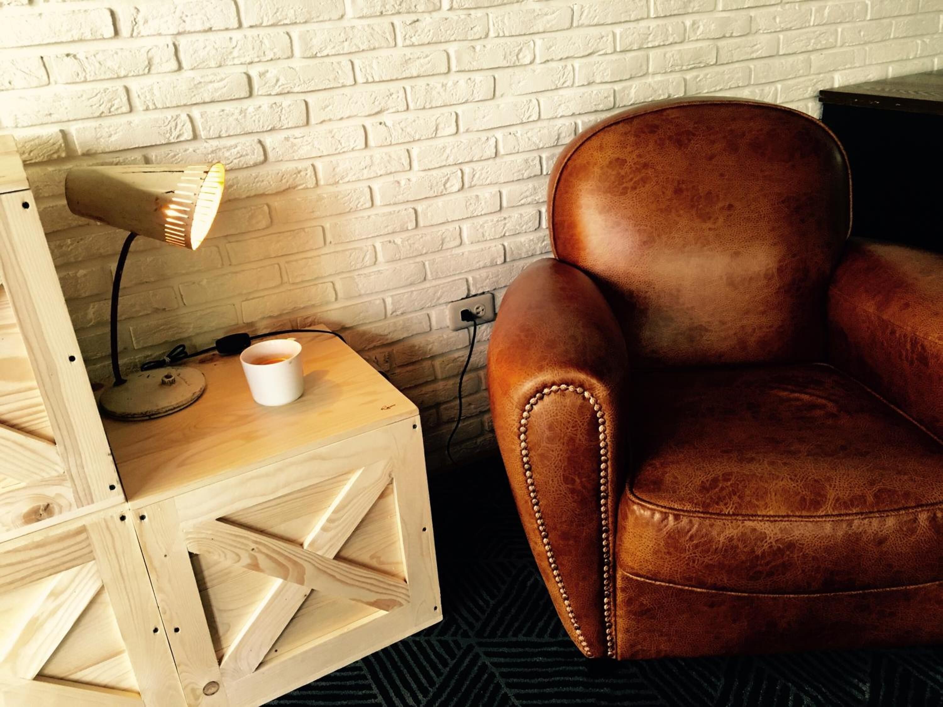 Een houten bijzettafeltje met een lamp en een witte mok, een bruine leren fauteuil, en een witte baksteenmuur op de achtergrond.