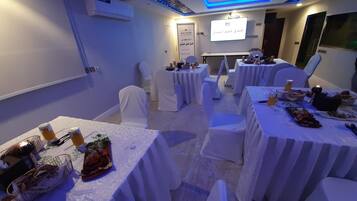Un salón de conferencias con mesas dispuestas para una reunión, una pantalla de proyector y un cartel con texto en árabe.