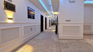 Een hotelgang met een marmeren vloer, witte wanden en een plafond met blauwe verlichting. Links bevinden zich deuren en rechts een lift.
