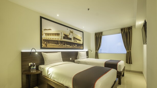 Quarto de hotel com duas camas, uma mesa de cabeceira com garrafas de água, um quadro na parede e uma janela com cortinas.