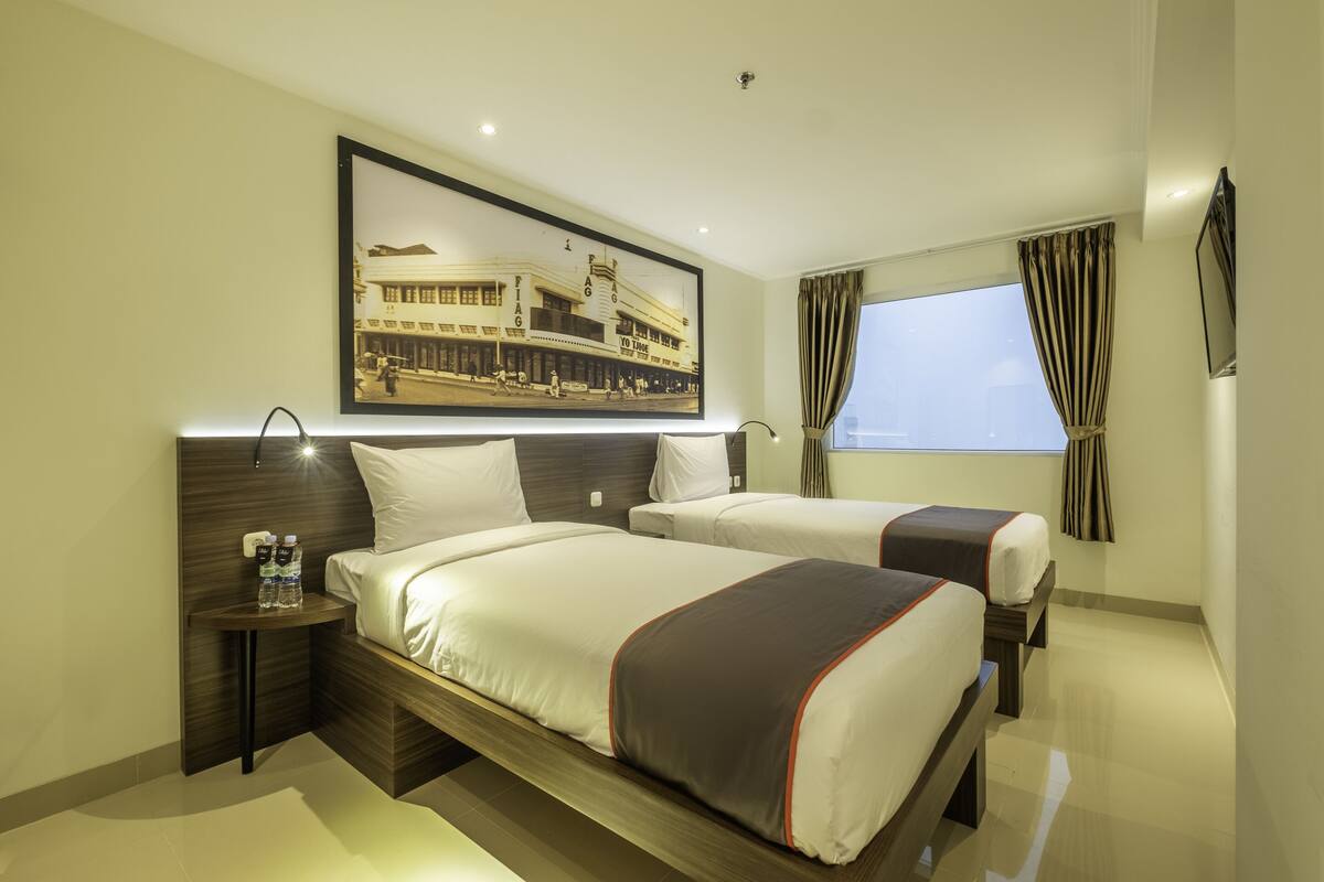 Habitación de hotel con dos camas, una mesita de noche con botellas de agua, un cuadro en la pared y una ventana con cortinas.