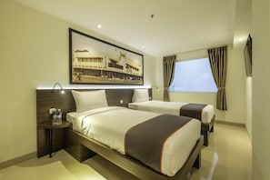 Quarto de hotel com duas camas, uma mesa de cabeceira com garrafas de água, um quadro na parede e uma janela com cortinas.