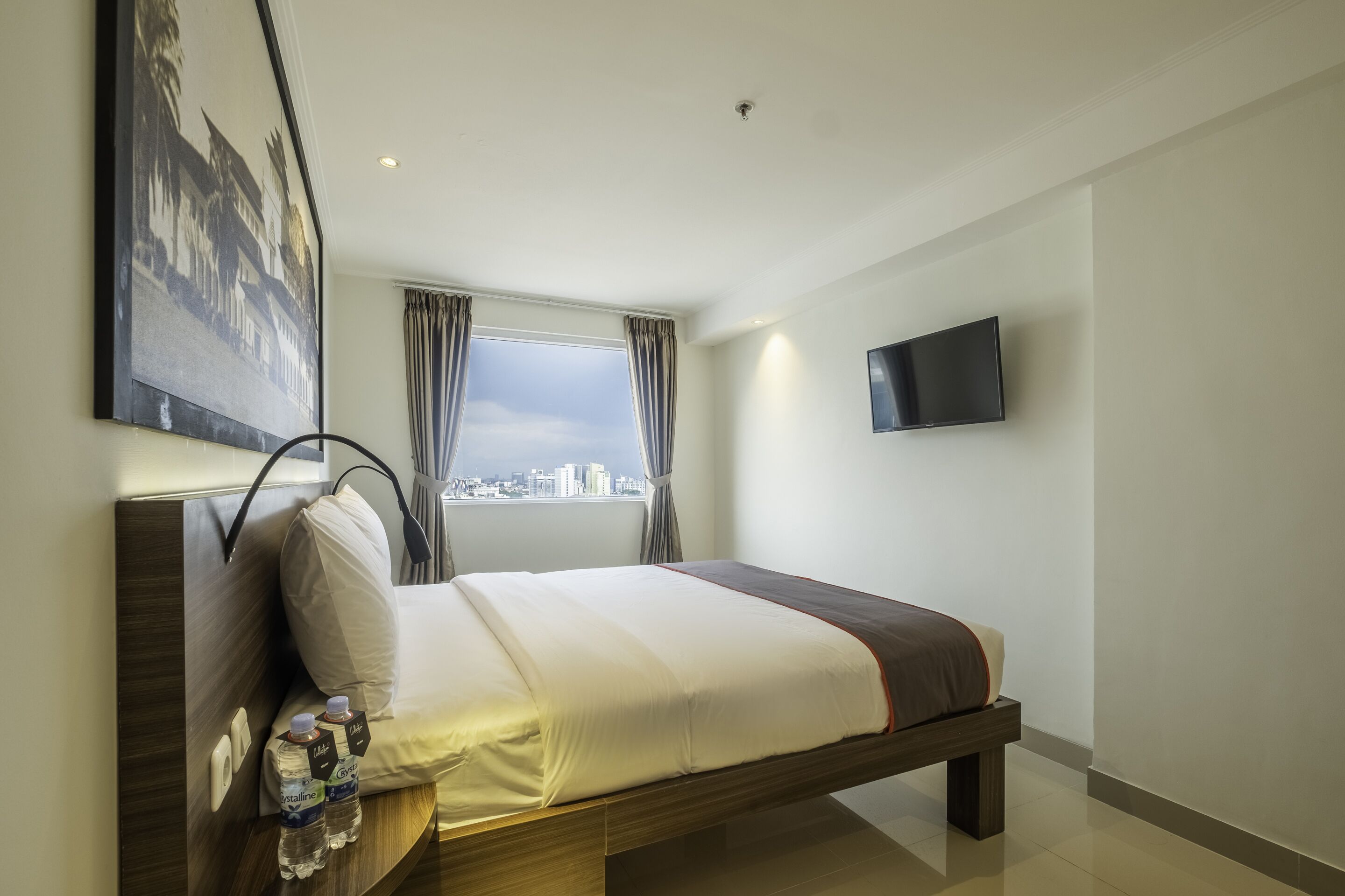 A modern hotel room with a large bed, a flat-screen TV mounted on the wall, and a cityscape view through the window.