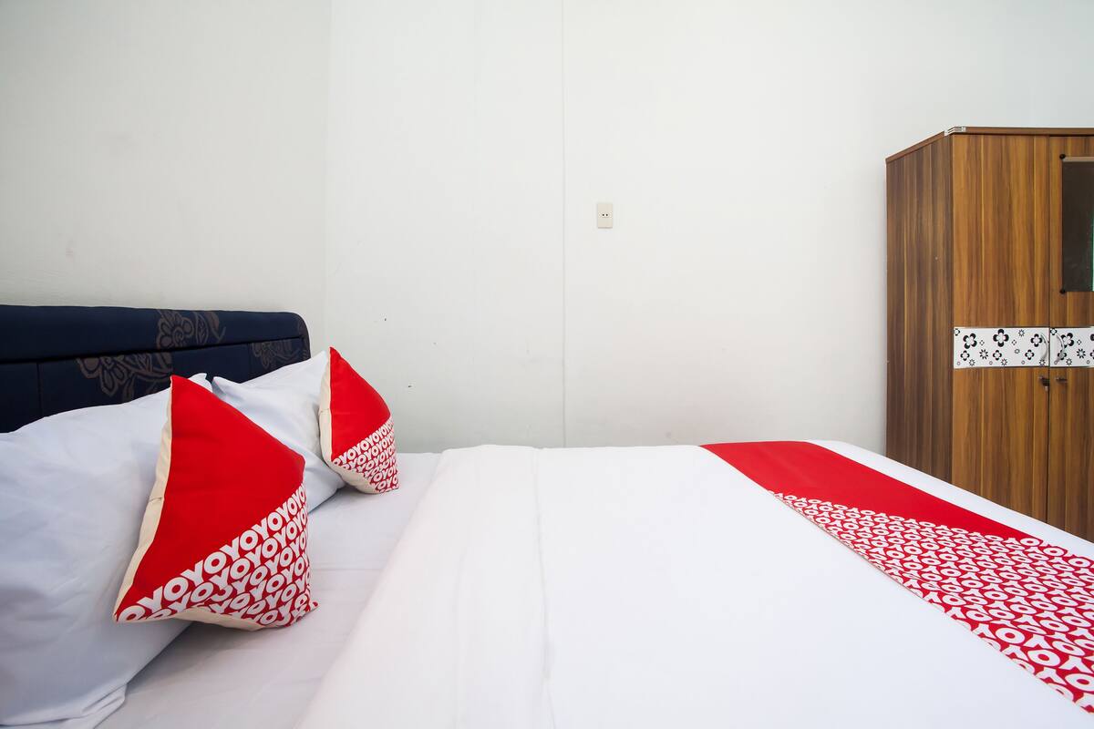 Un lit bien fait, avec une literie blanche et rouge, une armoire en bois et un mur blanc uni.