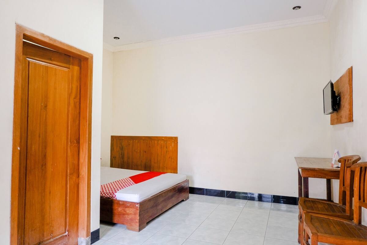 Una habitació amb un llit, escriptori, cadira, televisió penjada a la paret i una porta de fusta.