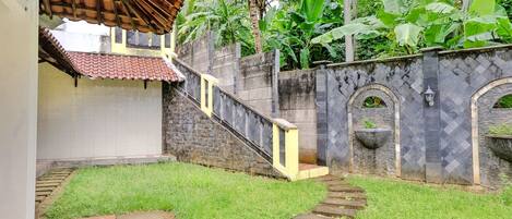 Un patio con una escalera de piedra que lleva a una zona cubierta, rodeado de abundante vegetación y un muro de piedra con arcos decorativos.