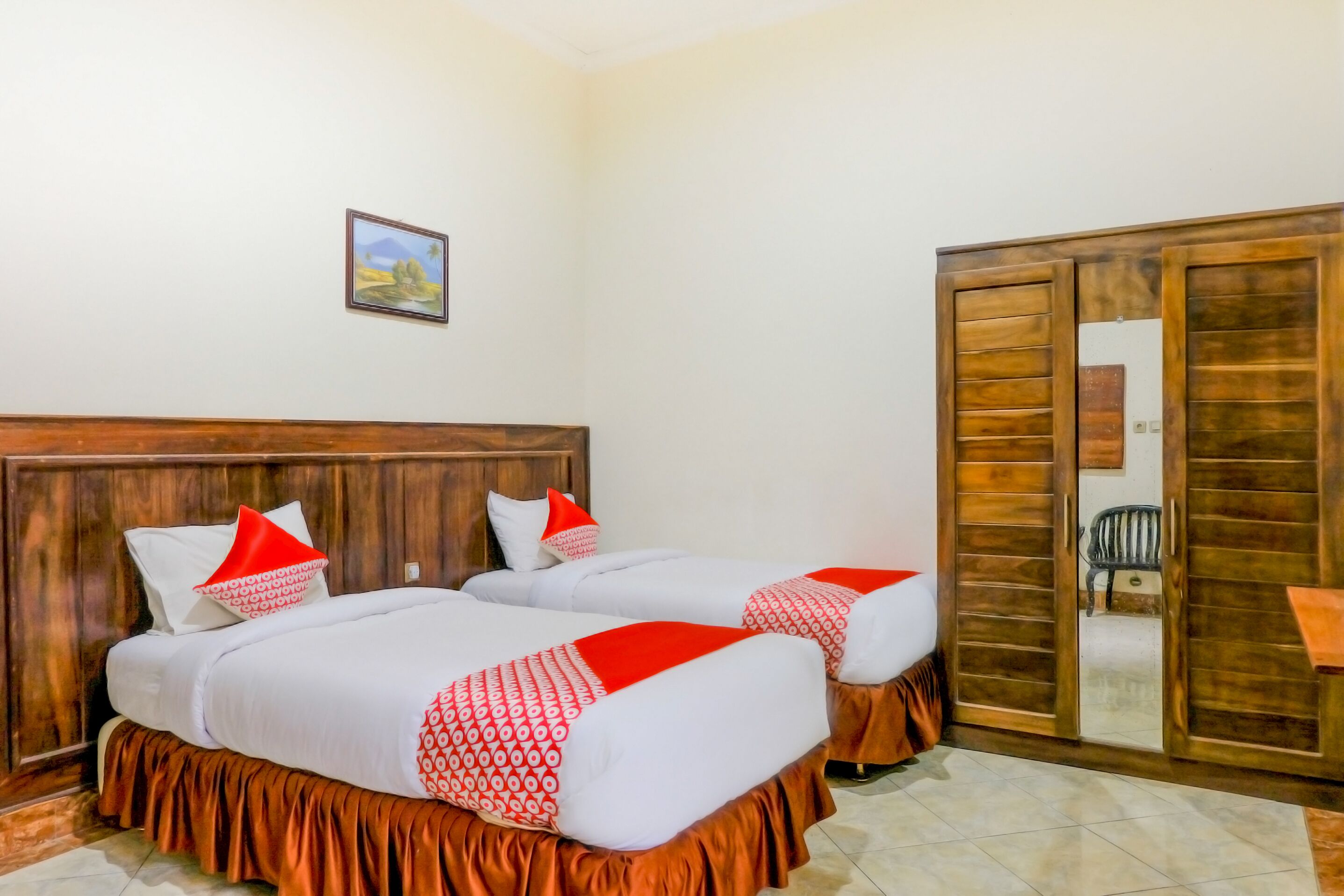 A room with two beds, each with a wooden headboard, white bedding, and red accents. There is a wooden wardrobe with a mirror and a small chair.