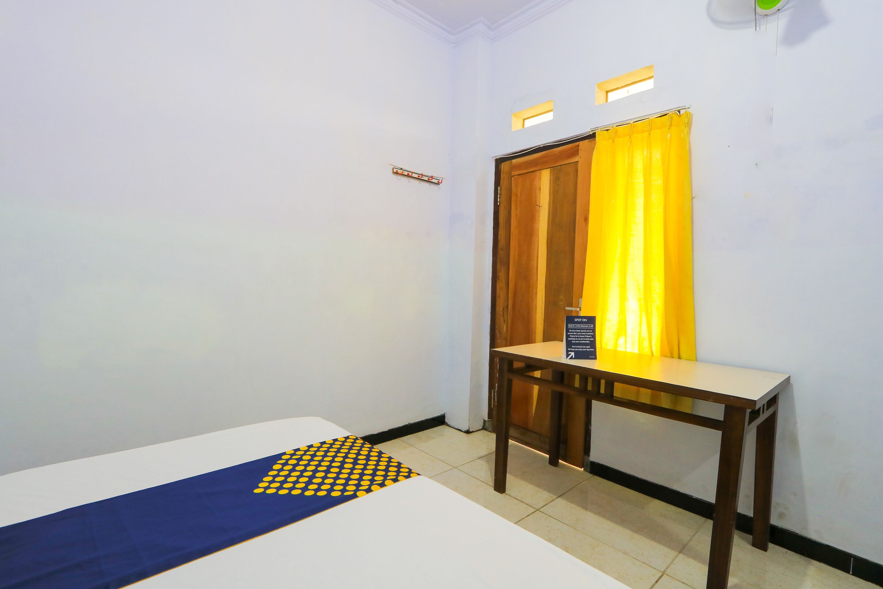 A small, clean room with a bed, a wooden table, and a yellow curtain.