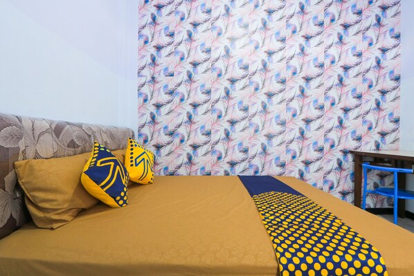 Un lit recouvert d’une couvre-lit à motifs jaunes et bleus, avec un oreiller marron et un oreiller bleu orné d’un motif jaune. On aperçoit au fond un papier peint à motifs représentant des plumes bleues et roses.