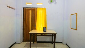 Een kamer met een houten deur, een geel gordijn, een tafel met een bordje, een ventilator aan de muur en een lijst met een spiegel.