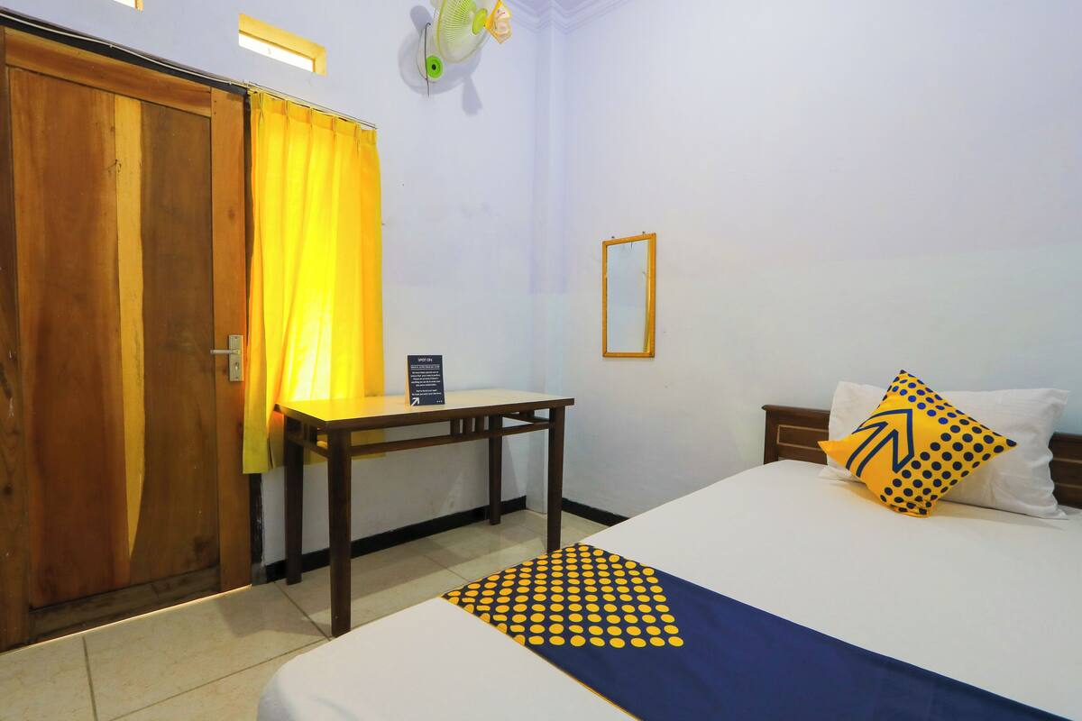 A hotel room with a bed, a desk, a chair, a wooden door, and a yellow curtain.
