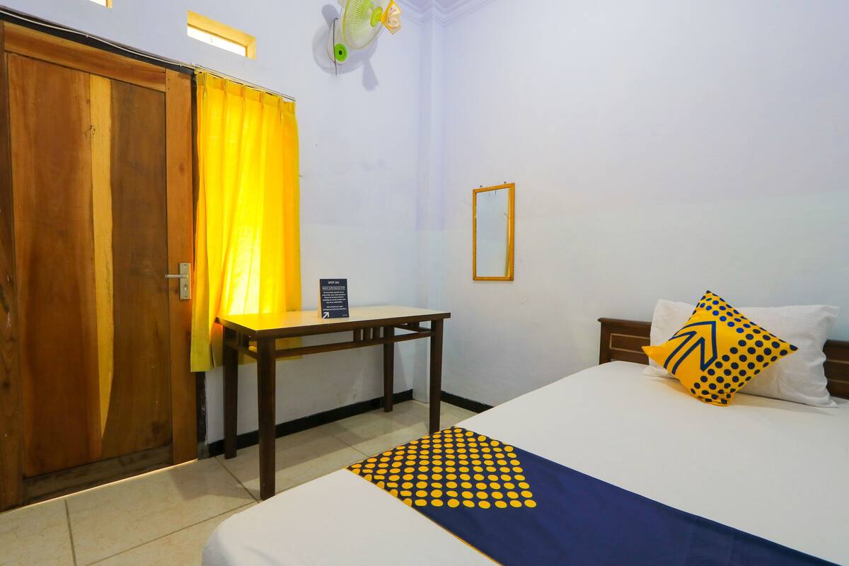 A hotel room with a bed, a desk, a chair, a wooden door, and a yellow curtain.
