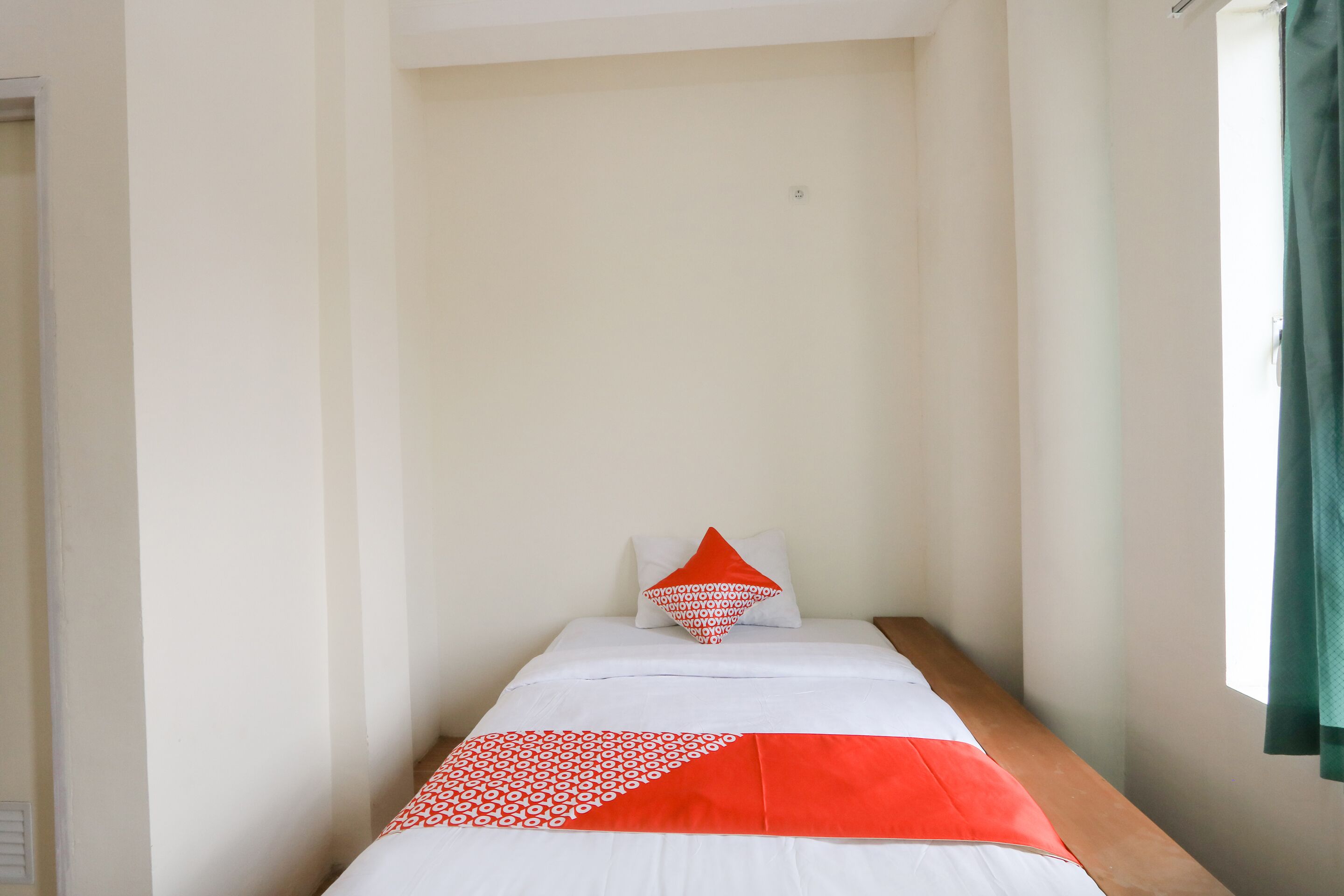 Una stanza piccola con un letto, una finestra con tenda verde e una parete bianca.